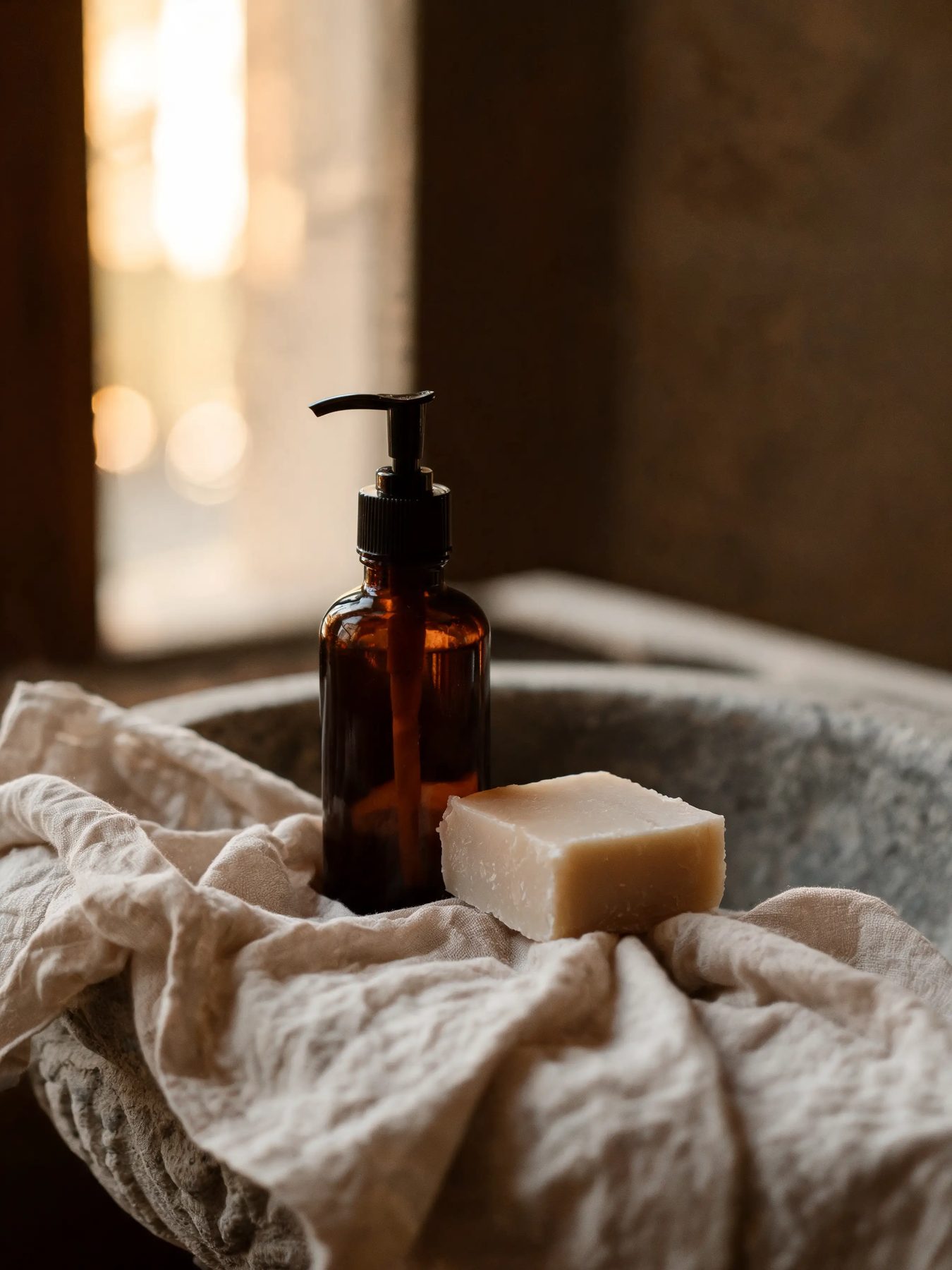Amber glass bottle and a cut bar of soap on linen beside a stone basin